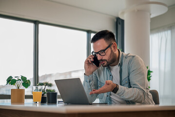 Obraz premium Worried businessman talking on cellphone while working on laptop at office