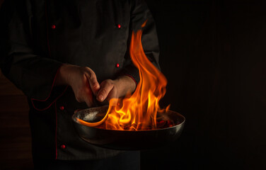 Professional chef cooking food in pan with fire flame on dark background. Restaurant and hotel service concept
