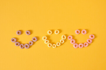 colored breakfast cereals laid out in the shape of a smiley face on a yellow background top view