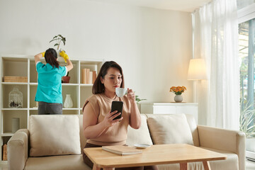 Serious woman drinking coffee and checking news on social media when cleaning service worker wiping dust from shelves in background