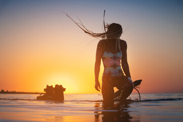 Silhouette frame. A young slim sexy female kitesurfer with a plank and a kiteboard stands in the water in the shallow water at sunset. Water sports. Stylized frame