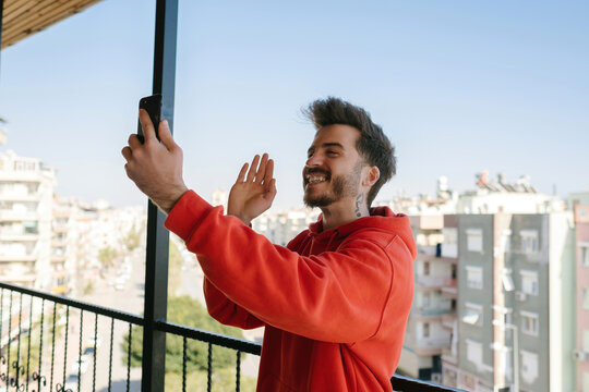 Young happy handsome man wearing red hoodie doing video call using smartphone at balcony. Waving hand video calling distance friend online in mobile chat app using smartphone videochat application.