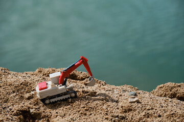Small plastic toy excavator with bucket working on sand extraction at quarry. Pond in background. Children's toy model of tractor extracts sand. Construction and mining industry. Creative banner.