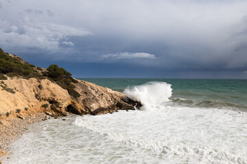 Stormy coastline with water splash and dark sky, sandy beach with turquoise sea, Sitges, Spain
