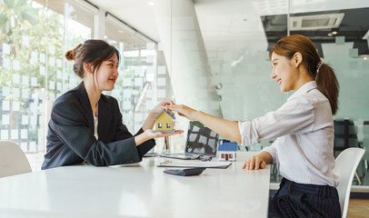 Female house agent sitting at the desk with house miniature and plan. Asian woman realtor giving contract information of new house to client.