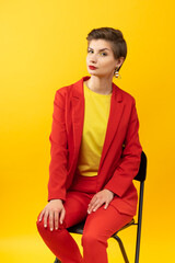 A model girl poses sitting on chair against a yellow background in the studio. Bright makeup, red jacket, yellow T-shirt, red trousers and yellow socks. Studio photoshoot business portrait