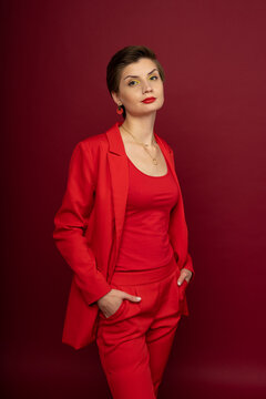 A woman in bright makeup, a red jacket, a red T-shirt, red trousers, with bright red lipstick and green shadows, stands on a red background with arms in pockets. Studio shooting