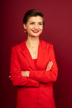 A boss business woman in bright makeup, a red jacket, a red T-shirt, with bright red lipstick and green shadows, stands on a red background with her arms crossed on her chest smiling. Studio shooting