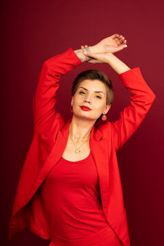 Gorgeous sexy seductive lady in a red (jacket, T-shirt. trousers), with a short haircut and bright makeup, posing in the studio on a red background with her arm uped
