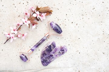 Facial massage kit - amethyst gua sha tool and face roller on stone concrete background with blooming sakura branch. Natural stone face massager, skin care concept, flat lay