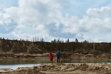 Sand quarry with water. Young couple in love is standing on sandy bank of river holding hands and looking into the distance. Travel with dog in a national park.