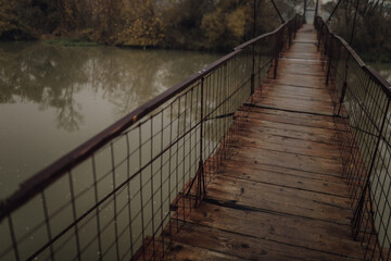 Eerie scenery of a wooden suspension bridge over the river in a cloudy day - obrazy, fototapety, plakaty