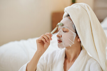 Fototapeta Woman closing eyes when massaging face with jade roller over brightening sheet mask obraz