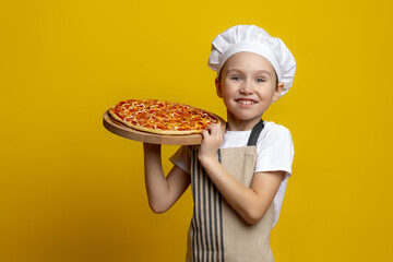  Cute 6-7-year-old boy in a chef's hat and apron holds a pizza tray. a little boy chef smiles sweetly with fast food in his hands on a yellow background. Copy space 