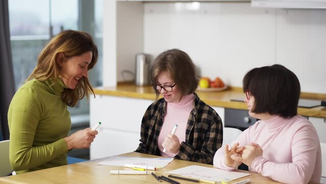 Two girls with down syndrome draw at the kitchen next to the teacher or mother together, slow motion
