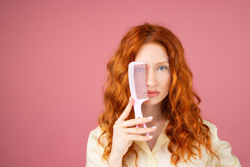 Close up photo of a redhead woman posing on pinkbackground covering one blue eye wioth a hair comb...