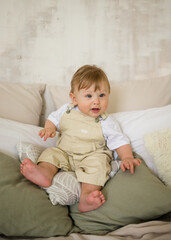 Caucasian baby boy in beige jumpsuit sits on pillows on the bed and looks at the camera