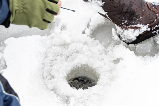 Fisherman sitting on frozen water near drilled hole in ice. Winter fishing concept. Hands in gloves holding small rod for catching fish