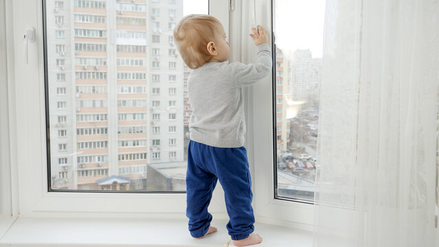 Little baby boy standing on windowsill and pulling window handle. Baby in danger. CHild safety.