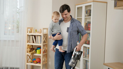 Young father holding his baby son while doing house cleanup and using vacuum cleaner.