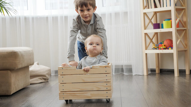 Older boy riding his baby brother in wooden toy cart in living room.
