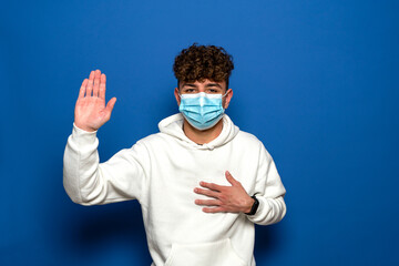 I swear. Portrait of serious honest Caucasian man with curly hair in surgical medical mask keeping hand on chest, palm up, promise, promise. indoor studio shot isolated on blue background