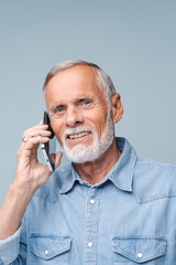 Joyful grandfather with grey beard talks to relatives on mobile phone smiling. Happy senior man in denim shirt enjoys conversation on blue background
