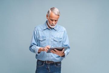 Grey-haired elderly man holds modern tablet checking mail. Bearded senior gentleman in elegant outfit stands on blue background using gadget studying online