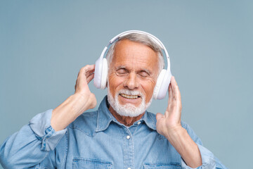 Fototapeta premium Senior man closes eyes listening to beautiful melody in headphones and smiles. Bearded gentleman enjoys sound standing on blue background closeup