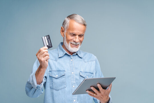 Elderly man makes online purchases on tablet holding credit card with hand. Bearded person in denim shirt stands on grey background looking at gadget