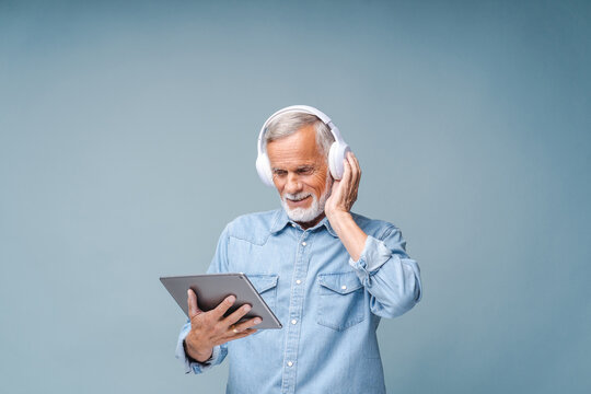 Senior man listens to educational audio in headphones holding modern tablet with hand. Bearded male person in denim shirt stands on blue background