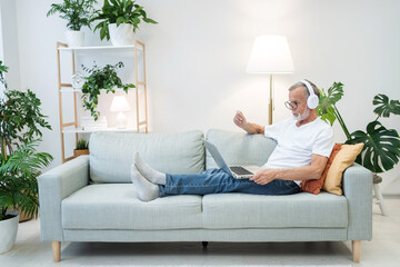 Senior man in headphones listens to educational audio using laptop at home. Elderly person in...