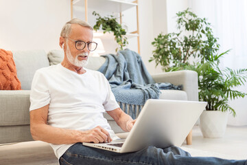 Bearded employee works online in laptop sitting on floor leaning on sofa. Focused senior man in glasses writes report after finishing task at home