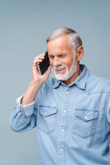 Bearded elderly man talks on mobile phone with friend smiling. Senior male person in denim jacket enjoys communication on blue background closeup