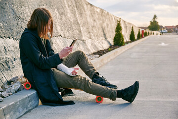 Fototapeta premium Teenager boy sitting on skateboard outdoor with trauma hand. Accidents and injuries to teenager during summer activities.
