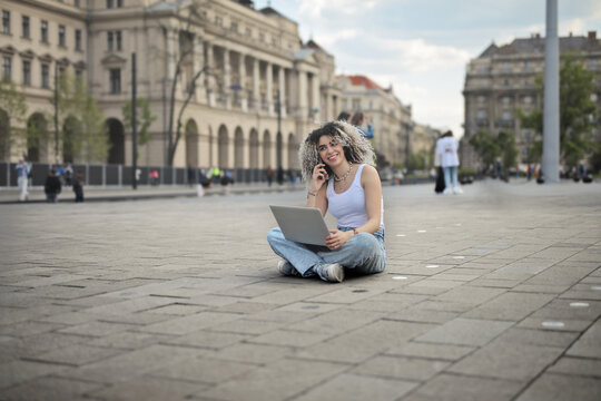 young woman in the street uses a laptop and talks on the phone