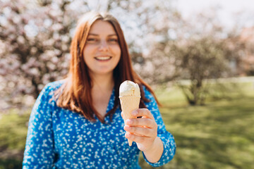 Happy young redhead woman with delicious white ice cream in waffle cone outdoors. Beautiful girl enjoying sweet food on nature background, summer time