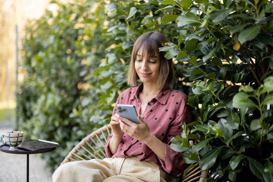 Young cheerful woman using smart phone while sitting on background of green bushes outdoors. Concept of online communication and spending time with gadgets outdoors