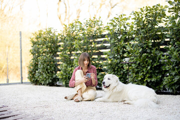Young woman sitting relaxed with her huge white adorable dog on green hedge background, spending...