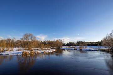 Fototapeta premium March sunny day by the river. A picturesque landscape, early spring, a river with snow-covered banks. The first thaws, the snow is melting.