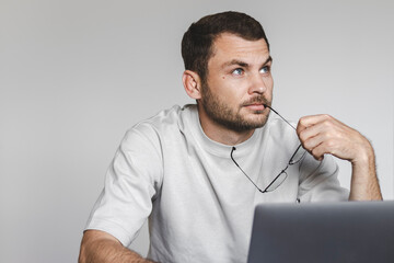Mid adult bearded man sits against a laptop holding eyeglasses in hand and looking up, dreaming,...