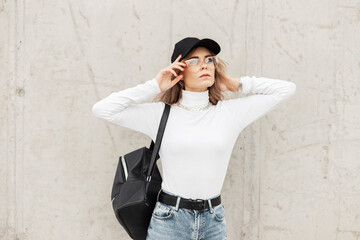 Stylish urban beautiful woman with glasses and a black cap in a fashionable white sweater with a backpack stands near a gray concrete wall in the city