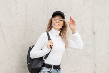 Happy beautiful young woman with a smile wearing fashionable urban clothes with a black cap, white sweatshirt and jeans with a backpack on a gray concrete background in the city