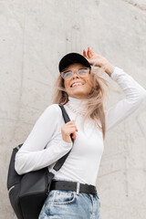 Happy beautiful woman with cute smile in casual clothes with black cap, white sweater, jeans, backpack and glasses walks and enjoys on the street near a concrete wall