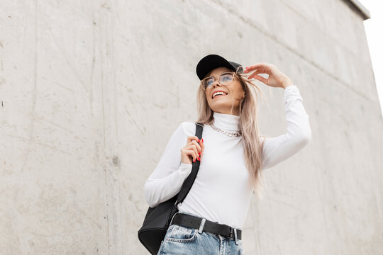Happy beautiful funny girl with a smile in fashionable casual clothes with a black cap and a backpack is walking on the street near a gray concrete wall