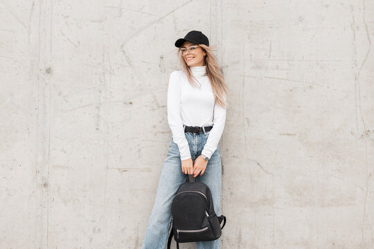 Beautiful young happy girl with a smile wearing fashionable clothes with a cap, white sweater, jeans and a backpack stands near a concrete gray wall