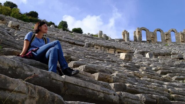 walk in Antalya Turkey on warm October afternoon. Pretty tourist woman with backpack at ruins of ancient city of Perge, large ancient amphitheater. resting after long walk on steps