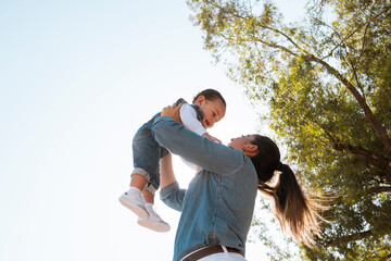 young mother lifts her son in her arms in the park at sunset - low angle