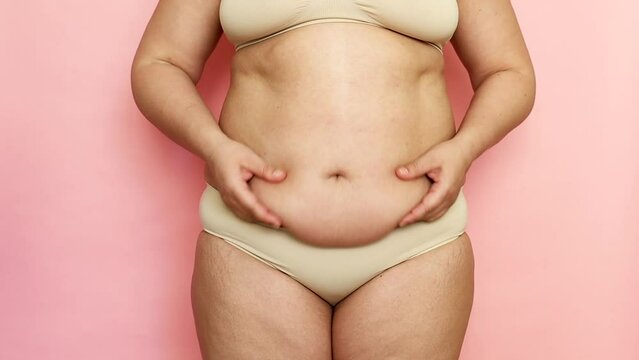 Unrecognizable fat and overweight woman holding folds of skin on thick large belly, obesity female body on pink isolated background, studio shot. Waving stomach. Get rid of visceral fat, weight gain
