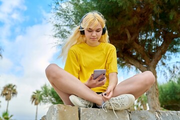 Naklejka na ściany i meble Beautiful female teenager in headphones with smartphone outdoor.
